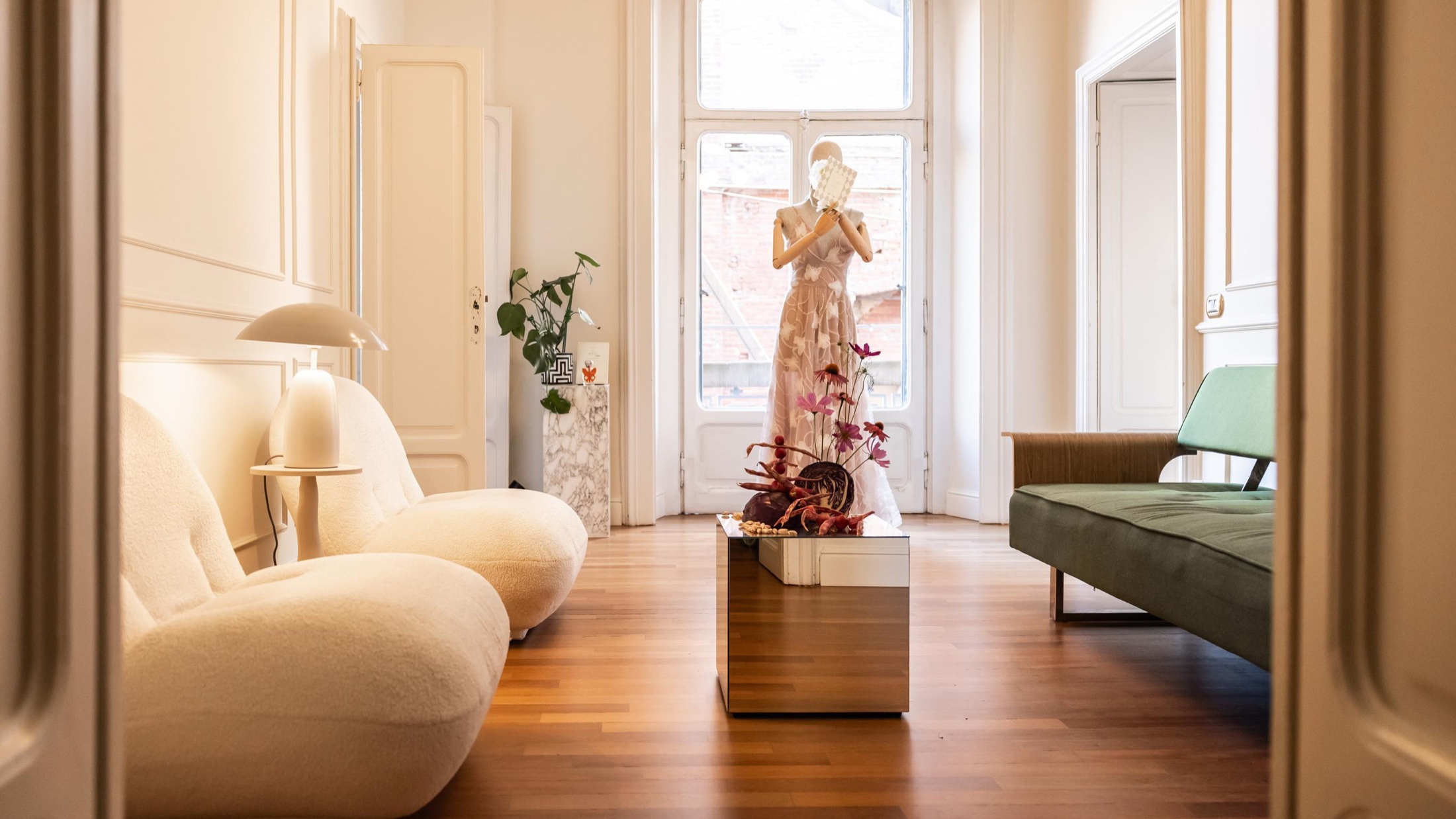 A view through the doorway into Salotto Duomo, with sculptural ivory armchairs, an olive sofa, wall panelling and a tall sunlit window